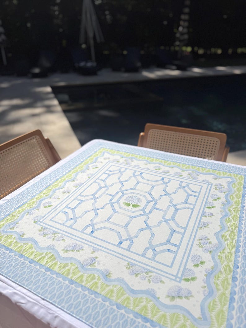 May include: A square tablecloth with a watercolor design. The central pattern features a blue geometric design with a floral motif. The border includes light blue, green, and white floral and geometric patterns. The tablecloth is set on a table with two wicker chairs.