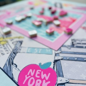 May include: A close-up of a New York-themed board game. The game features a pink apple with the text "NEW YORK" and a map of the city. Small, colorful game pieces are scattered across the board. The background includes illustrations of cityscapes and luggage.