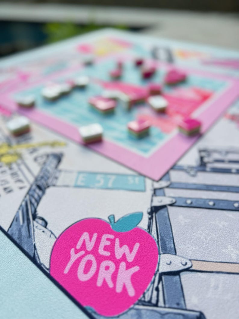 May include: A close-up of a New York-themed board game. The game features a pink apple with the text "NEW YORK" and a map of the city. Small, colorful game pieces are scattered across the board. The background includes illustrations of cityscapes and luggage.