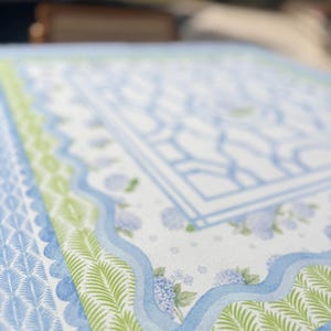 May include: Close-up of a patterned fabric featuring a light blue and white design with a central rectangular motif. The border includes green fern-like patterns and a scalloped edge. Hydrangea floral accents are also visible.