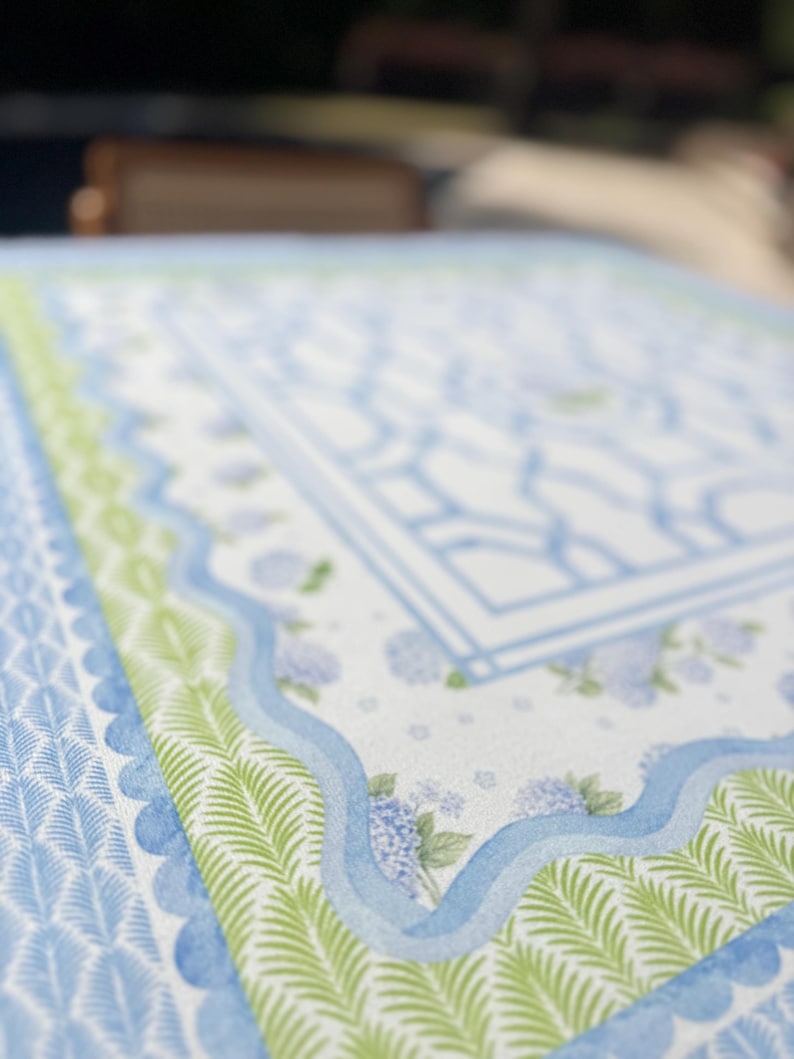 May include: Close-up of a patterned fabric featuring a light blue and white design with a central rectangular motif. The border includes green fern-like patterns and a scalloped edge. Hydrangea floral accents are also visible.