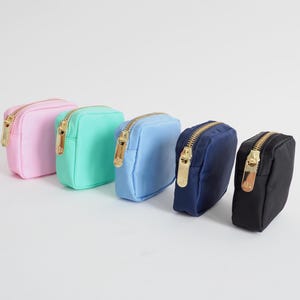 May include: Five small, square cosmetic bags in a row, each with a gold zipper. The bags are in pastel colors: pink, mint green, light blue, navy blue, and black. The bags are made of a smooth, solid-colored material.