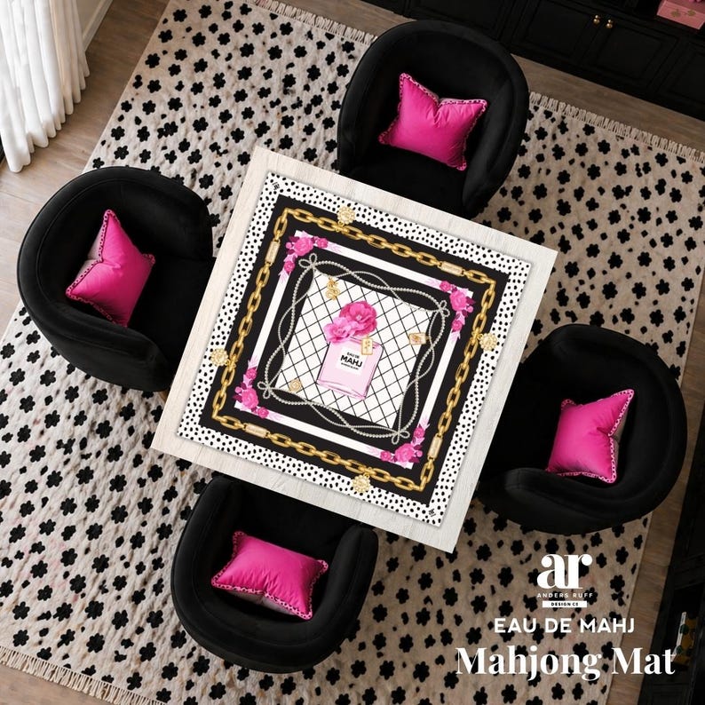 May include: A square white Mahjong mat with a perfume bottle design, surrounded by black, gold, and pink accents. The mat is centered on a black and white patterned rug, with four black armchairs and pink pillows.
