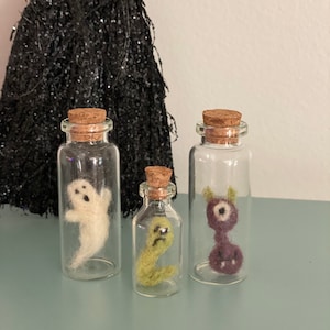 Puede incluir: Tres botellas de vidrio con tapas de corcho, cada una contiene una criatura de Halloween de fieltro diferente. Las botellas est&aacute;n sobre una superficie azul claro. Las criaturas son un fantasma blanco, un gusano verde y un monstruo morado con un ojo.