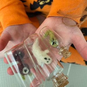 Puede incluir: Tres peque&ntilde;as botellas de vidrio con tapones de corcho, cada una contiene una criatura de Halloween de fieltro diferente. Las botellas est&aacute;n en la mano de una persona. Las criaturas son un gato negro, un fantasma blanco y un monstruo verde.