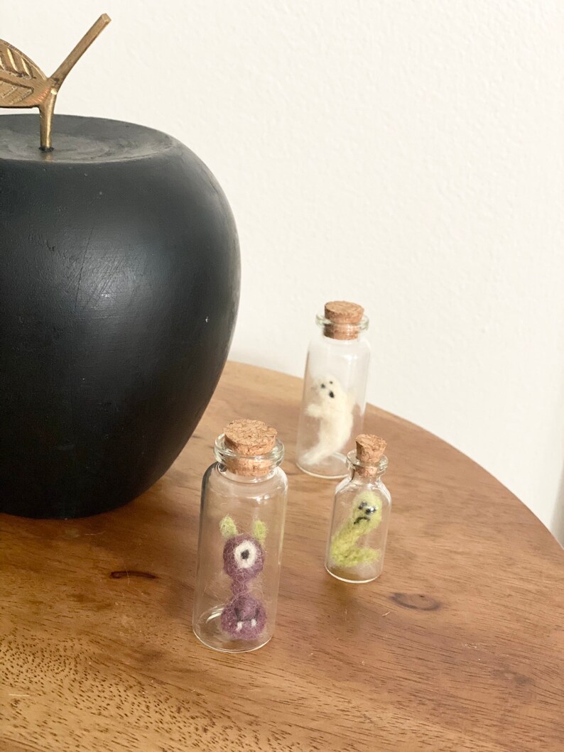 Puede incluir: Tres peque&ntilde;as botellas de vidrio con tapas de corcho, cada una contiene una criatura de fieltro diferente. Las criaturas son un monstruo morado, un gusano verde y un fantasma blanco.
