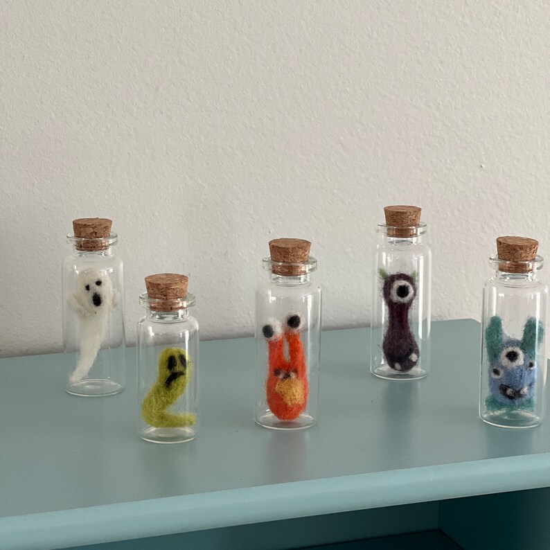 Puede incluir: Cinco peque&ntilde;os frascos de vidrio con tapas de corcho, cada uno contiene una criatura de fieltro diferente. Las criaturas son un fantasma, un gusano, una criatura roja y naranja con ojos, una criatura morada con un ojo y una criatura azul con un ojo.