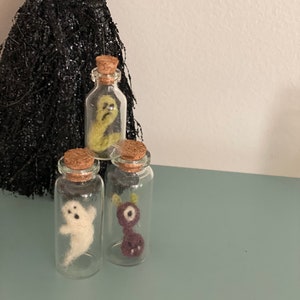 Puede incluir: Tres peque&ntilde;as botellas de vidrio con tapas de corcho, cada una contiene una figura de fantasma de fieltro diferente. Los fantasmas son verde, blanco y morado.