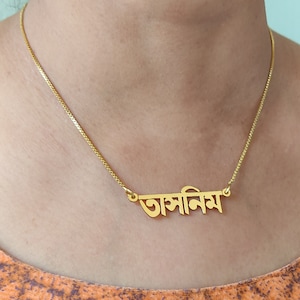 Puede incluir: Un collar de cadena de oro con un colgante de placa con la palabra bengalí "আসিমা" en oro.
