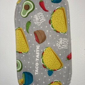 May include: A gray fabric kitchen towel with a white border and a repeating pattern of yellow tacos, green avocados, red peppers, and the text "Hola Taco!" and "Taco-Tastic!"