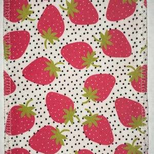 May include: White fabric with a repeating pattern of red strawberries and black polka dots. The fabric has a stitched edge.