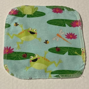 May include: Square, light blue fabric with a repeating pattern of green frogs, lily pads, pink flowers, bees, and a dragonfly. The edges are finished with a white zigzag stitch.