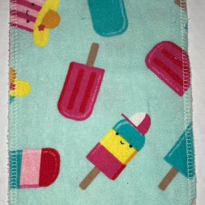May include: A turquoise blue fabric square with a white stitched border. The fabric has a repeating pattern of colorful popsicles with different designs. Some popsicles have a smiling face, others have stripes, and some have a watermelon design.