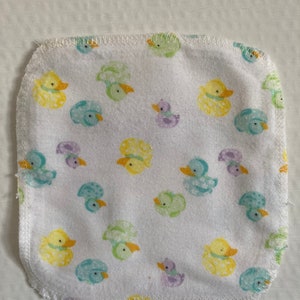 Può includere: Un quadrato di tessuto bianco con un motivo di anatre gialle, verdi e viola. Il tessuto ha uno sfondo bianco con un bordo cucito.