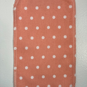 May include: A peach-colored fabric square with white polka dots. The fabric has a white stitched border.