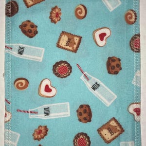 Puede incluir: Una tela azul con un borde cosido blanco presenta un patrón repetitivo de botellas de leche, galletas y galletas con chispas de chocolate. La tela tiene un fondo azul claro con un borde cosido blanco.