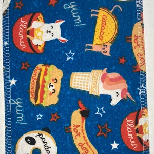 May include: A blue fabric with a white border and a variety of colorful illustrations of food and animals. The illustrations include a hot dog, a hamburger, a donut, a unicorn, a llama, and a taco. The text "yum!" appears multiple times on the fabric.