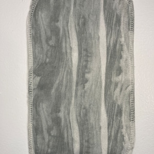 May include: A grey fabric burp cloth with a white stitched border. The fabric has a wood grain pattern.