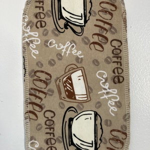 Puede incluir: Una toalla de cocina de tela marrón con un diseño de taza de café y granos de café. La toalla tiene la palabra "café" escrita varias veces.