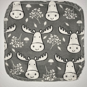 May include: A gray fabric square with a white stitched border. The fabric has a repeating pattern of white moose with black antlers and black eyes, and white berries and mushrooms.