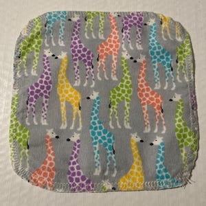 Puede incluir: Un paño de eructo cuadrado gris para bebé con un patrón repetido de jirafas coloridas. Las jirafas son en tonos de morado, amarillo, naranja, verde y azul con manchas blancas. Los bordes están acabados con una puntada en zigzag blanca.