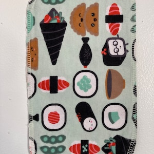 Puede incluir: Paño rectangular verde claro con un patrón repetido de ilustraciones de sushi. Los diseños de sushi incluyen nigiri, rollos maki y temaki, en tonos de rojo, marrón, negro y verde. El paño tiene un borde cosido blanco.