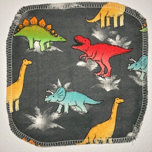 May include: A dark gray fabric square with a white stitched border features a repeating pattern of colorful dinosaurs. The dinosaurs include a green stegosaurus, a red Tyrannosaurus Rex, a blue Triceratops, and two yellow brachiosaurs.