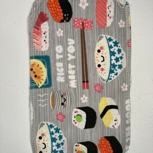Puede incluir: Tela rectangular gris con un patrón de sushi. El diseño incluye varios tipos de sushi, palillos y el texto "Nice to Meet You". Los bordes están acabados con una costura blanca.