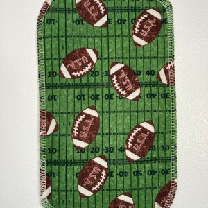 May include: A green fabric square with a white stitched border. The fabric has a pattern of brown footballs with the text "U.S.A." on them. The footballs are on a green and white grid pattern that resembles a football field.