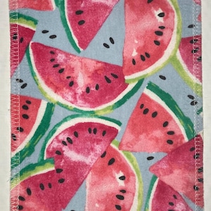 May include: Rectangular fabric with a watermelon pattern. The design features slices of watermelon in shades of red and pink, with black seeds and green rinds, set against a light blue background. The edges are finished with white stitching.