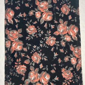 Puede incluir: Un parche de tela negro con un patrón floral. La tela tiene un diseño floral rosa y blanco sobre un fondo negro. El parche tiene una puntada en zigzag alrededor de los bordes.