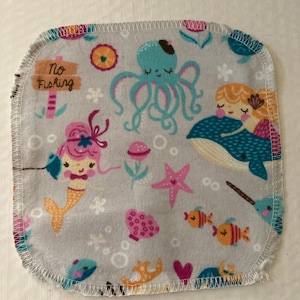 May include: A gray, square fabric with a whimsical underwater scene. The design includes mermaids, an octopus, fish, and a sign that reads "No Fishing." The edges are finished with a white, stitched border.