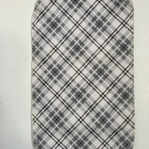 Puede incluir: Un parche de tela blanca con un patrón de cuadros negros y grises. La tela está cosida alrededor de los bordes con hilo blanco.