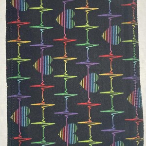 Puede incluir: Tela rectangular negra con un patrón repetido de formas de corazón de arcoíris y líneas de electrocardiograma. Los corazones están rayados verticalmente con los colores del arcoíris. Los bordes están cosidos con hilo a juego.