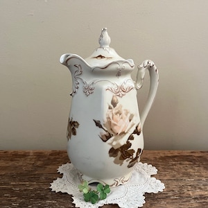 May include: White porcelain creamer with a floral design featuring brown and pink roses. The creamer has a lid and a handle.