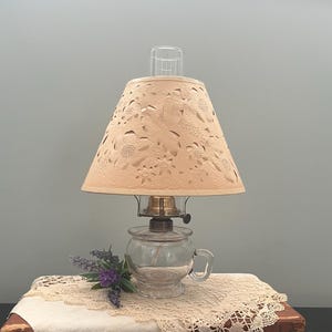 May include: A vintage oil lamp with a beige, sunflower-patterned lampshade. The lamp sits on a small wooden table covered with a cream-colored lace doily and a white cloth. A small bouquet of purple flowers adds a touch of color.