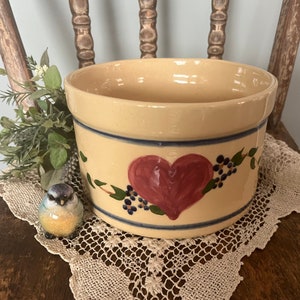 May include: A yellow ceramic crock with a hand-painted red heart design and blue and green floral accents. The crock is sitting on a white crocheted doily.