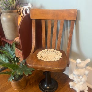 May include: A wooden chair with a slatted back and a round, dark metal pedestal base. A circular, cream-colored crocheted doily rests on the flat wooden seat. The chair features a warm, medium-brown wood finish with visible grain and a sturdy construction.