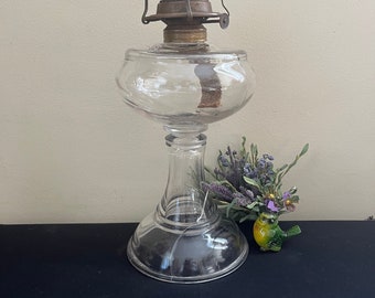 May include: A clear glass oil lamp with a metal burner and a round base. The lamp is on a black surface with a small green and yellow bird figurine and a bunch of purple flowers.