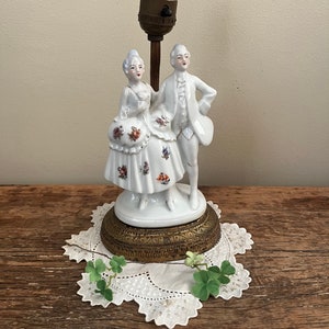 May include: A white porcelain lamp base featuring a couple in 18th-century attire. The woman holds a bouquet of flowers and the man has his hand on his hip. The lamp base has a gold-colored metal base with intricate details.