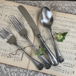 May include: A set of silverware with ornate handles, including a knife, fork, and spoon, laid out on a sheet of music with the text "Studies from "Morning Prayer." 125"