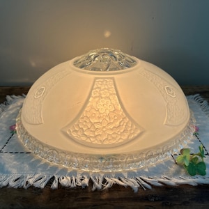 May include: A vintage, frosted glass lampshade with a floral design. The shade has a rounded shape with a clear glass decorative top and a detailed floral pattern. The lampshade is resting on a white fringed cloth.