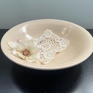 May include: A beige ceramic bowl with a white lace doily and a white flower with a gold center.