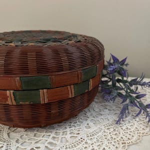 May include: A round, woven basket with a lid. The basket is primarily brown with bands of green and red. The basket sits on a white lace doily, with purple flowers in the background. The basket appears to be vintage.