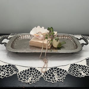 May include: A silver rectangular tray with handles, holding a seashell, small books, a decorative bird, and a pearl necklace. The tray sits on a white lace doily with a black table in the background.