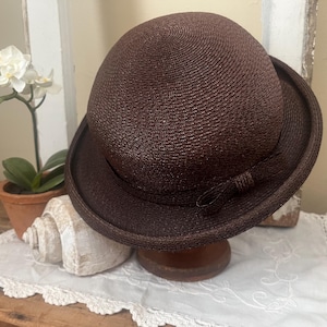 May include: A dark brown cloche hat with a rounded crown and a small brim. The hat has a woven texture and a small bow detail on the side. It is displayed on a wooden stand, with a seashell and a potted plant in the background.