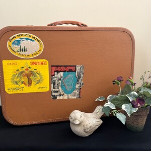 May include: A brown vintage suitcase with several stickers, including one that says "SEMIRAMIS CAIRO" and another that says "GRAND NEW HOTEL NAZARET", sitting on a black surface next to a small potted plant with purple flowers and a white ceramic bird.