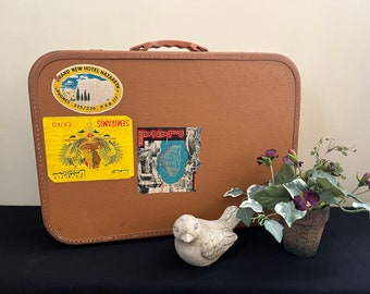 May include: A brown vintage suitcase with several stickers, including one that says "SEMIRAMIS CAIRO" and another that says "GRAND NEW HOTEL NAZARET", sitting on a black surface next to a small potted plant with purple flowers and a white ceramic bird.