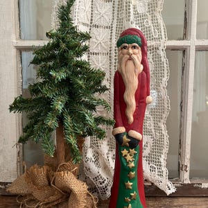 May include: A carved Santa Claus figurine in a red robe with a long white beard and a green hat. The figurine holds a green stocking adorned with gold stars. A small artificial Christmas tree sits to the left, wrapped in burlap.