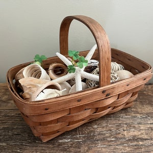 May include: A brown woven basket with a handle filled with white seashells, a white starfish, and green leaves.
